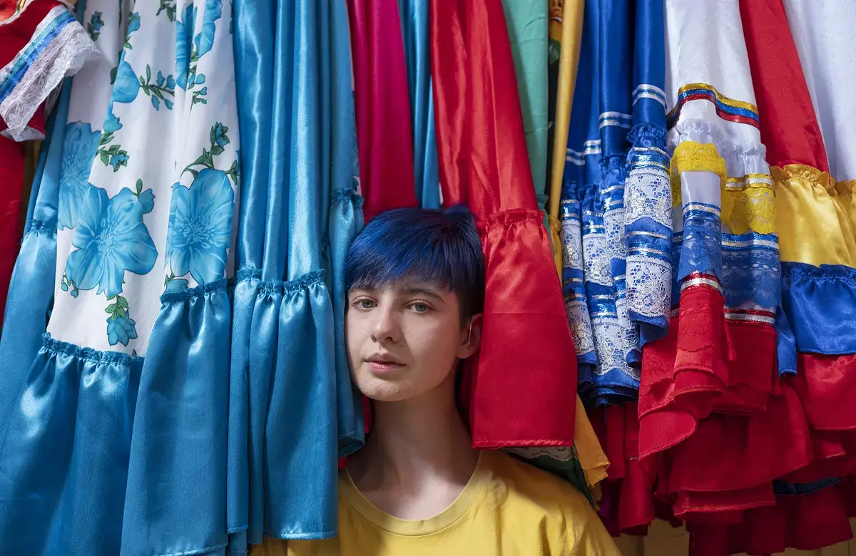 A young person with blue hair sits at the hems of a row of brightly colored dresses