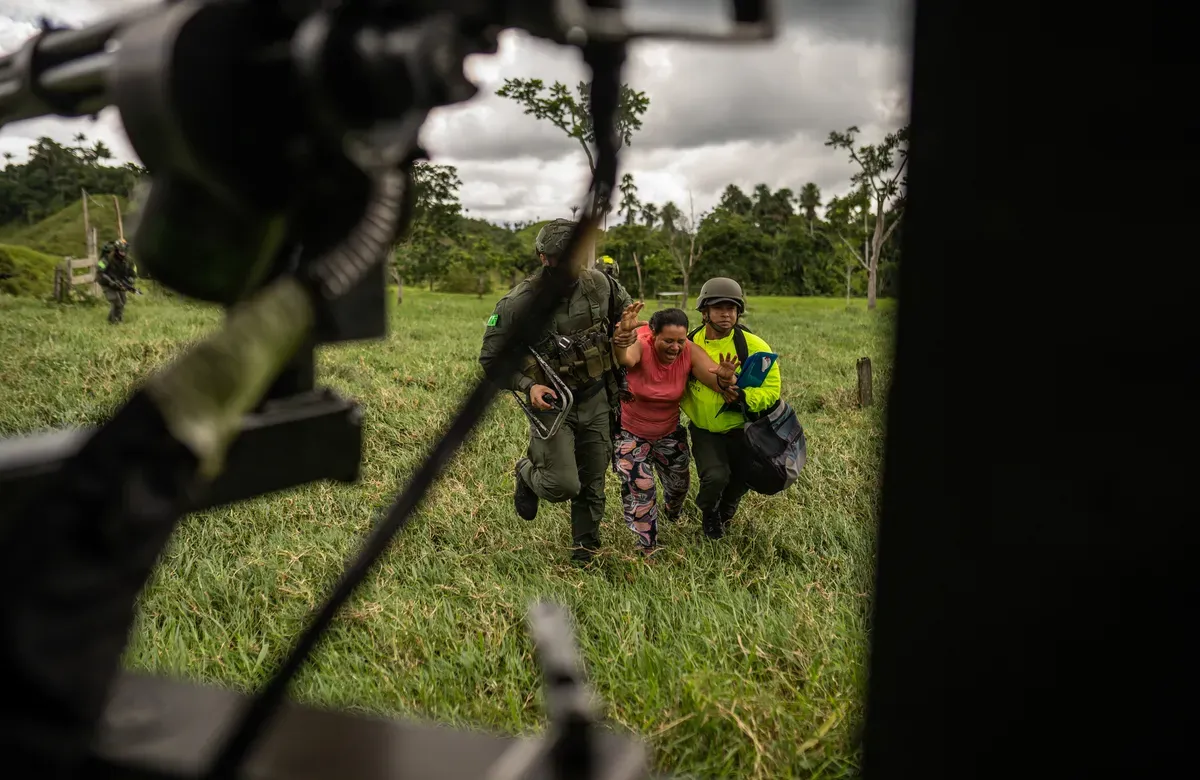 framed through a helicopter, a woman is being forcibly carried to the vehicle by two soldiers