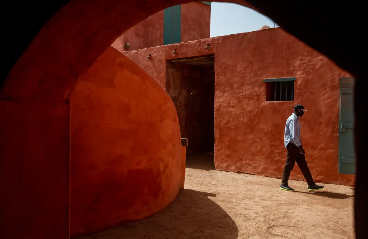 against the framing of an darkened archway, a man wearing a face mask walks down an alleyway against red walls