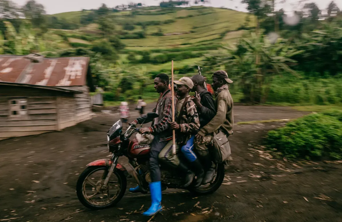 A group of four men ride in a moving motorcycle holding various weapons against a green landscape
