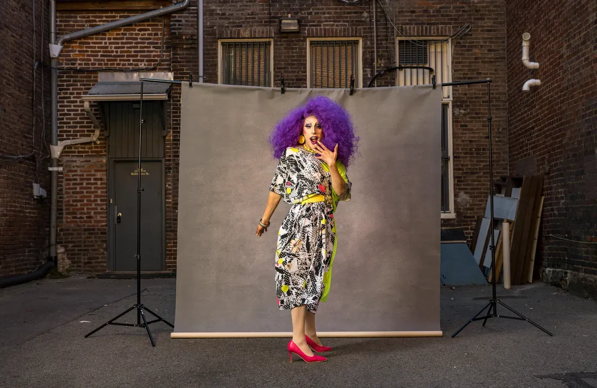 A drag queen with purple hair poses in front of a backdrop in an alley