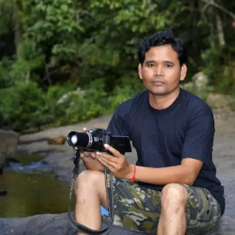 leng_ouch.jpg Portrait of Leng Ouch holding a camera and sitting on a rock next to a lake with trees behind him.
