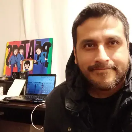 A person wears a black jacket. In the foreground, there is a study desk with a laptop and a poster board.