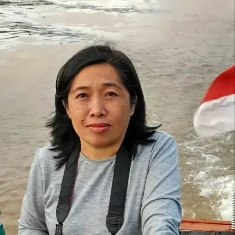 A person wears a grey long sleeve shirt and is carrying a camera. In the foreground there is a beach with ocean waves.