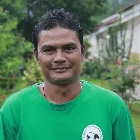 Image of a person wearing a green shirt. A house and tropical forest is in the background.