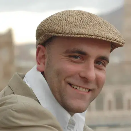 A man wears a brown hat, white scarf, and a brown jacket.
