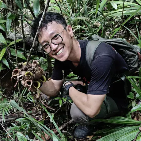 A man wears a black shirt and trekking pants. He has eyeglasses and is holding a plant.