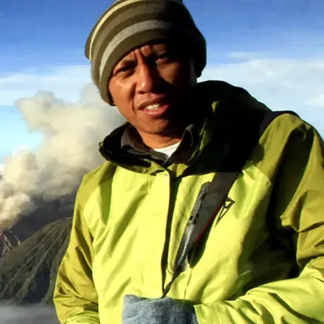 A man wearing a beanie and a yellow jacket. In the background, there is a mountain range and fog.