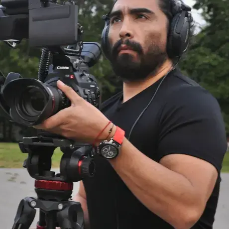 enrique.jpeg Enrique Huaiquil holds a camera that is set up on a tripod. He wears headphones and focuses on the camera.