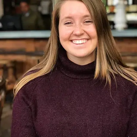 A woman with long blonde hair wearing a purple sweatshirt looks at the camera