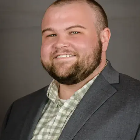 A man with short hair wears a gray jacket and flannel shirt.
