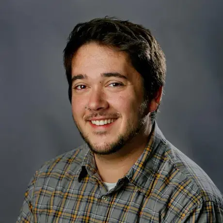 A man with short dark hair wears a plaid button down against a dark background