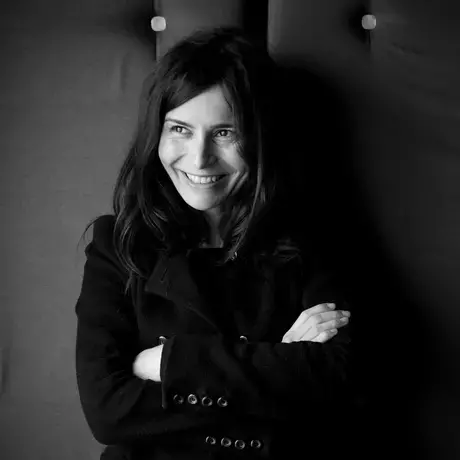 A black and white photo of a woman with long dark hair sitting down with her arms crossed, wearing a black jacket