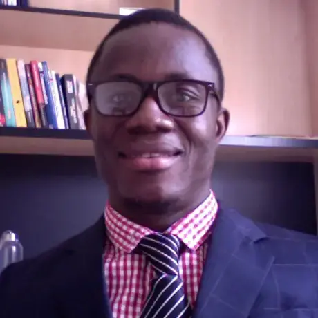 A man with dark skin, short hair and glasses smiles, with a bookshelf in the background