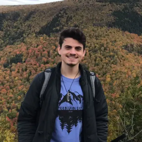Man stands in black jacket and blue shirt in front of forest