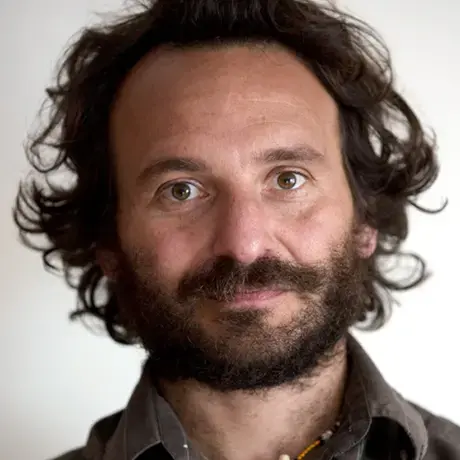 A man with curly brown hair and facial hair wears a brown button down