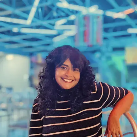 A woman with dark curly hair wearing a striped shirt against a bright blue-lit background