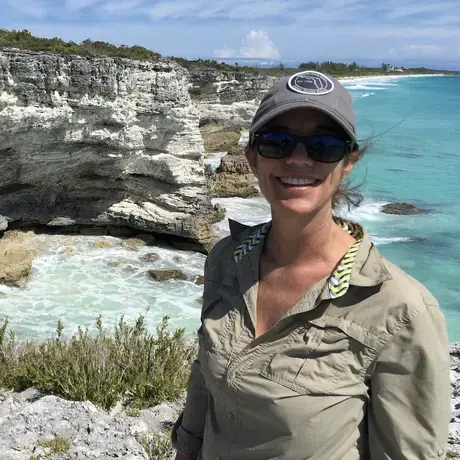 A woman wearing a baseball cap and green button-down against a background of cliffs beside the ocean