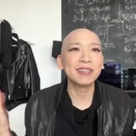 A person with a bald head wearing a leather jacket sits at a desk and gestures with both hands