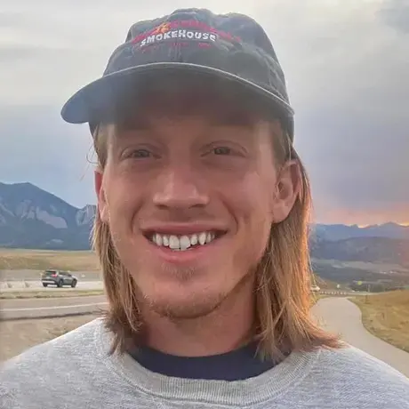 A man with shoulder-length blond hair wearing a dark blue baseball cap and grey sweatshirt against a grassy landscape