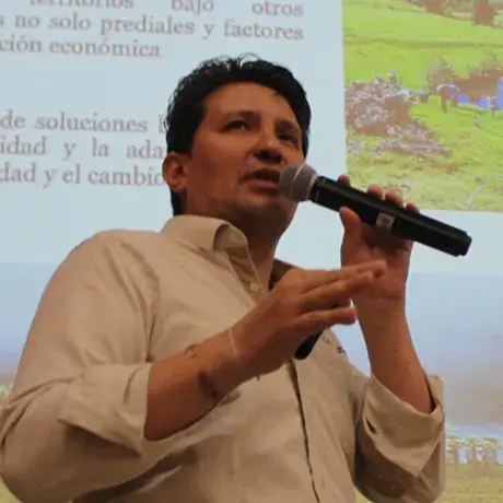 A man wearing a light button-down shirt holds and speaks into a microphone with slides projected behind him