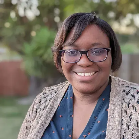 A smiling woman with short dark hair and glasses wearing a blue shirt and tan cardigan