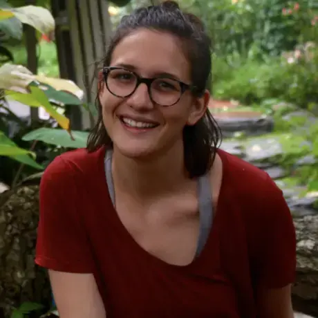 A woman with dark shoulder-length hair and glasses wearing a red t-shirt against a leafy background