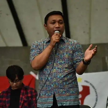 A man in a short sleeve button down speaks into a microphone on a stage