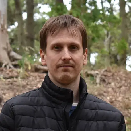 A man with short brown hair wearing a black puffer coat in a wooded area