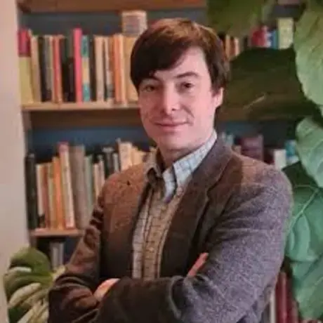 A man with short brown hair wearing a button down shirt and brown jacket stands against a bookshelf folding his arms