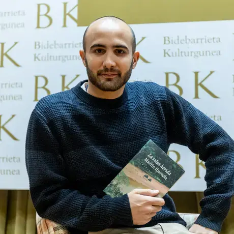 A man wearing a blue shirt sits holding a book in his arms.