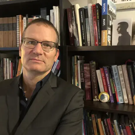 A man wearing glasses and a grey blazer against a bookshelf
