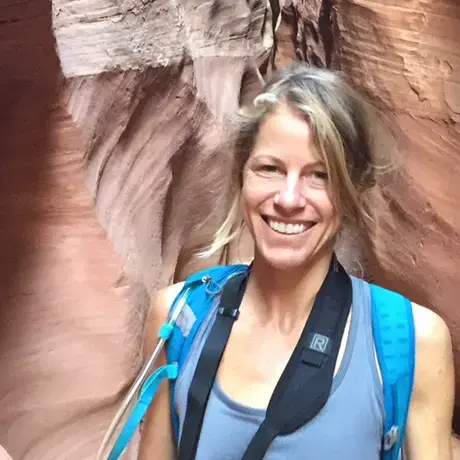 Carlyle wears a blue shirt and backpack while walking through a rocky formation