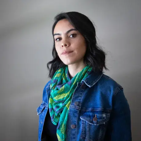 Headshot of Greta wearing a jean jacket and green scarf against a grey background