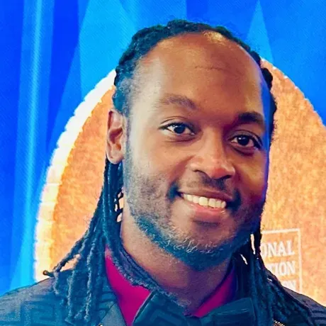 Kenyon Andrews is a Black man with a beard and dreadlocks pulled back from his face. He wears a suit and smiles at the camera.