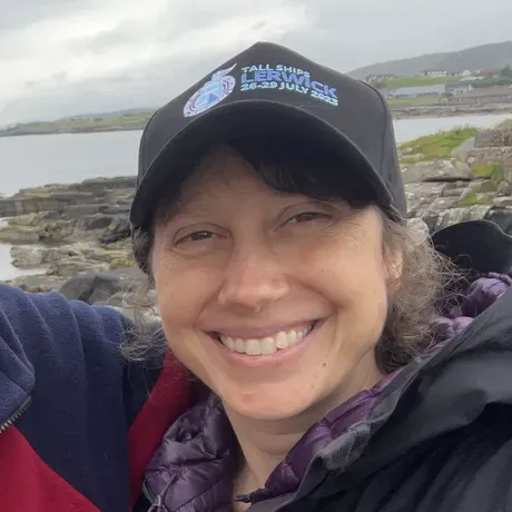 Susan Greenwald is a white woman wearing a jacket and athletic cap. She is smiling at the camera.