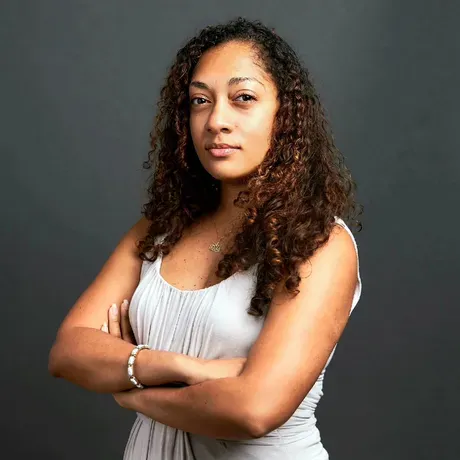 Lucia is a woman of color wth arms folded in a commanding stance looking directly into the camera
