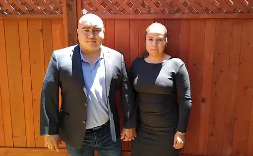 Jose Jaime &amp; Gabriela Covarrubias, parents of Christian Madrigal, are standing together in front of a wooden fence.