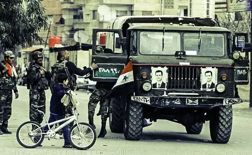 A young boy walks with a bicycle past Syrian army soldiers in the city of Harasta, 9 km north of Damascus. Photo courtesy of Freedom House flickr. Syria, 2012.