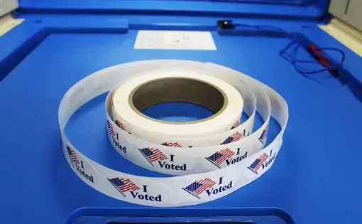 Red white and blue "I voted" stickers with American flag are on a spool of stickers placed on a voting booth platform. Image by&nbsp;Barbara Kalbfleisch. United States, undated.&nbsp;
