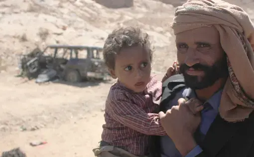 Mabkhout Ali al Ameri stands with his 18-month-old son, Mohammed, in the village of al Ghayil in Yemen’s al Bayda province. Mabkhout’s wife, Fatim Saleh Mohsen, was shot in the back of the head by helicopter gunship fire as she fled with Mohammed in her arms during a U.S. raid. Image courtesy of The Intercept. Yemen, 2017.