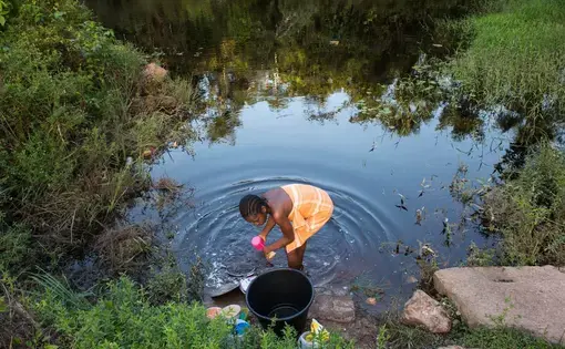 Osyanda Misidjan, 17, washes dishes on the banks of the Coermotibo River in her village of Adjuma Kondre, Suriname. Once surrounded by rainforest, the green palm trees and valley soccer field of the village are now surrounded by stony, bauxite-red plains left over from Alcoa's mining operations that ceased in 2015. Image by Stephanie Strasburg. Suriname, 2017.