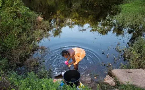 Osyanda Misidjan, 17, washes dishes on the banks of the Coermotibo River in her village of Adjuma Kondre, Suriname. Once surrounded by rainforest, the green palm trees and valley soccer field of the village are now surrounded by stony, bauxite-red plains left over from Alcoa's mining operations that ceased in 2015. Image by Stephanie Strasburg. Suriname, 2017.