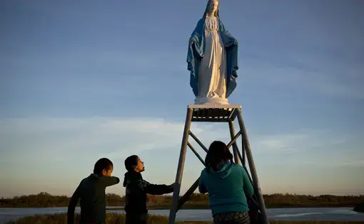 Young people play at a statue of the Virgin Mary near the St. François-Xavier Catholic Church on the shores of the Attawapiskat River in northern Ontario. The community has a complicated history with the Catholic Church, with many of its members subjected to horrible abuses while at the Catholic-run St. Anne’s Residential School. Image by David Maurice Smith/Oculi. Canada, 2016.