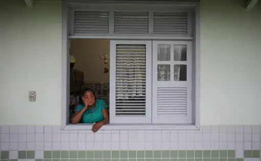 Maria da Silva Trindade peeks out of her living room window to chat with the other residents of Marituba’s former leprosy colony. Trindade was diagnosed with the disease at the age of 15, and she moved into the colony the same year. Nearly 80 years later she’s still there. Image by Anton L. Delgado. Brazil, 2020.