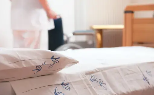 Bed at nursing home with nurse and wheelchair in background. Image by Stokkete / Shutterstock.com. Undated.