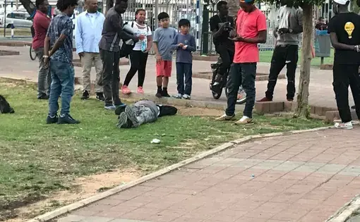 A Sudanese refugee reacts to substance overdose and bystanders surround him in Levinsky Park in south Tel Aviv, Israel. Many refugees suffer various mental health disorders such as post-traumatic stress disorder (PTSD), anxiety, depression, alcohol and substance abuse, and in extreme cases, psychosis. Image by Isma'il Kushkush. Israel, 2018.