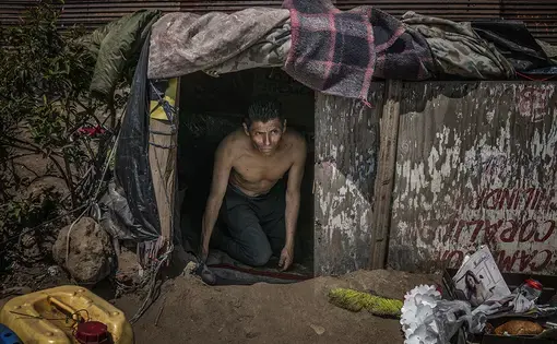 Luis—a deportee from the Mexican State of Querétaro, emerges from his makeshift shelter high atop the first desolate hilltop mesa east of the Pacific Ocean beside the U.S./Mexico border wall.  Image by James Whitlow Delano. Mexico, 2017.