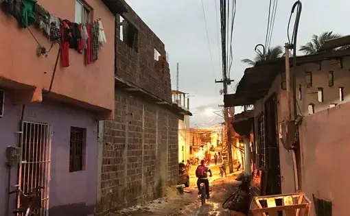 Image of a narrow street at dusk in a community on the outskirts of Recife, Pernambuco. There is an unpaved dirt road with small homes packed close together on either side. A teenager sits on a stoop in the shadows, while a young boy can be seen riding his bike down the street. Image by Poonam Daryani. Brazil, 2017.