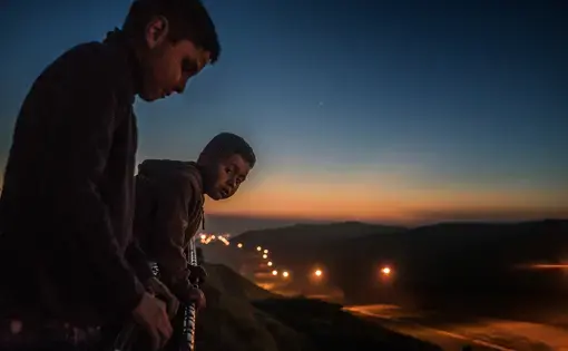 Young boys from the Arias family peer over the first, older US-built border wall at dusk into the flood-lit no-man's land created between the two walls/fences where US Border Patrol is present 24 hours a day, seven days a week.  Colonia Libertad, Tijuana, Mexico.  The Arias family is led by Estel, these boys’ grandmother and matriarch of the clan.  Their house actually rests up against the border wall and yet the family expressed no fear from human or drug traffickers.  Still their home, despite being…